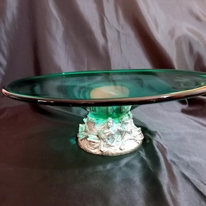 May include: A decorative cake stand with a round, emerald green glass top and a silver-colored base. The base features sculpted figures. The stand is designed to display cakes, pastries, or other desserts.