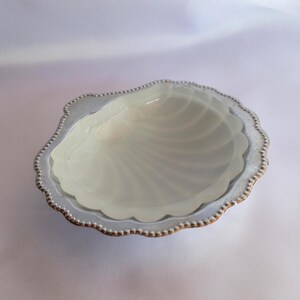 Könnte beinhalten: Eine dekorative, cremefarbene Schale in Muschelform mit gewelltem Rand. Der Rand ist mit einer silberfarbenen Zierleiste und kleinen goldfarbenen Perlen umrandet. Das Innere der Schale hat ein perlmuttartiges Finish.