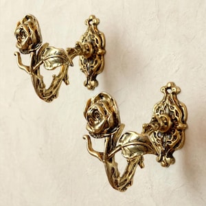 May include: Two wall-mounted hooks crafted from a metallic gold-toned material. Each hook features a three-dimensional rose blossom at the end of a stem with a single leaf, attached to an ornate, elongated oval backplate with intricate detailing.