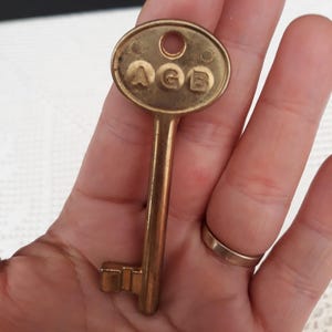 Puede incluir: Una llave de latón antigua con las letras "AGE" estampadas en la cabeza ovalada. La llave tiene un vástago largo y delgado y una paleta rectangular. La llave se sostiene en una mano, mostrando el tamaño y los detalles.
