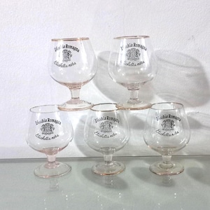 Puede incluir: Cinco copas de brandy transparentes con bordes y tallos dorados. Cada copa presenta el texto "Vecchia Romagna Etichetta Nera" en negro. Las copas están dispuestas sobre una superficie reflectante.