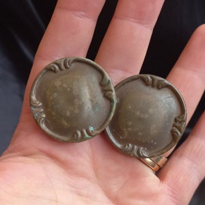 Puede incluir: Dos botones antiguos de color latón con adornos de volutas en relieve en los bordes. Los botones redondos muestran un aspecto envejecido, con signos de edad y oxidación verdosa. Cada botón mide aproximadamente 3 cm de diámetro.