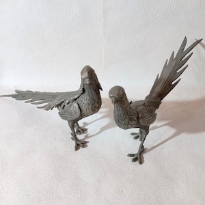 Könnte beinhalten: Zwei antike Metallvogel-Figuren. Die Vögel sind mit Federn detailliert und stehen auf zarten Beinen. Ein Vogel hat seine Schwanzfedern ausgebreitet, während der andere seine Schwanzfedern gefaltet hat. Die Vögel sind von gedämpfter Silberfarbe.