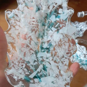 K&ouml;nnte beinhalten: Ein klares, blattf&ouml;rmiges Dekorationsteil, gef&uuml;llt mit wei&szlig;en und t&uuml;rkis farbenen Fragmenten. Die Kanten des Teils sind glatt und wellenf&ouml;rmig. Das Objekt wird in einer Hand gehalten, mit einem unscharfen Hintergrund.