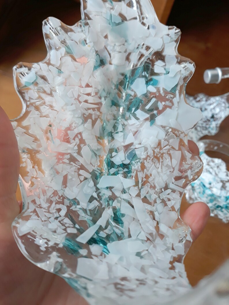K&ouml;nnte beinhalten: Ein klares, blattf&ouml;rmiges Dekorationsteil, gef&uuml;llt mit wei&szlig;en und t&uuml;rkis farbenen Fragmenten. Die Kanten des Teils sind glatt und wellenf&ouml;rmig. Das Objekt wird in einer Hand gehalten, mit einem unscharfen Hintergrund.