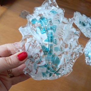 K&ouml;nnte beinhalten: Ein dekoratives Objekt in Form einer Muschel aus klarem Glas mit wei&szlig;en und t&uuml;rkisfarbenen Akzenten. Das Glas hat ein strukturiertes, fragmentiertes Aussehen mit einem gewellten Rand. Das Design beinhaltet eine zentrale, gerippte Struktur mit t&uuml;rkisfarbenen Highlights.