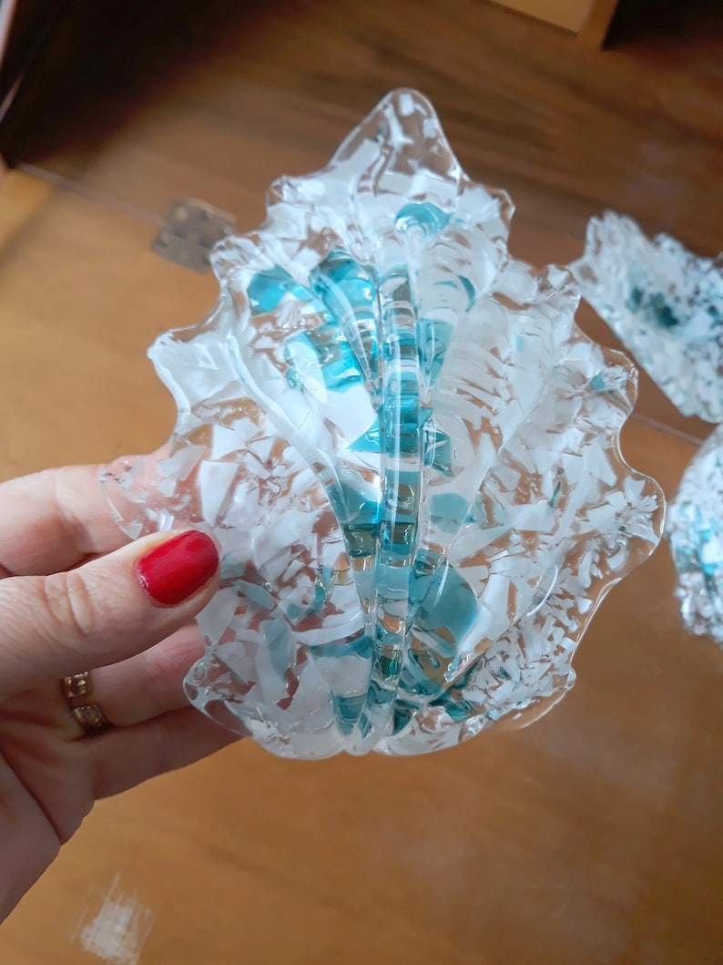 K&ouml;nnte beinhalten: Ein dekoratives Objekt in Form einer Muschel aus klarem Glas mit wei&szlig;en und t&uuml;rkisfarbenen Akzenten. Das Glas hat ein strukturiertes, fragmentiertes Aussehen mit einem gewellten Rand. Das Design beinhaltet eine zentrale, gerippte Struktur mit t&uuml;rkisfarbenen Highlights.