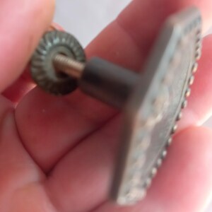 May include: A metallic cabinet knob with a hexagonal face featuring a beaded border. The knob has a central post and a threaded metal screw extending from the back, accompanied by a small, circular, ridged metal washer for secure installation.