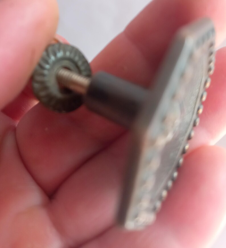 May include: A metallic cabinet knob with a hexagonal face featuring a beaded border. The knob has a central post and a threaded metal screw extending from the back, accompanied by a small, circular, ridged metal washer for secure installation.