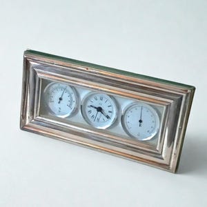 Könnte beinhalten: Eine rechteckige Wetterstation in Silberoptik mit drei runden Messgeräten. Das mittlere Messgerät zeigt eine Uhr mit schwarzen Zeigern und römischen Ziffern. Die beiden anderen Messgeräte haben weiße Zifferblätter mit schwarzen Markierungen und Nadeln. Der Rahmen hat eine grüne Rückseite.