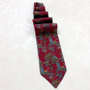May include: A red necktie with a repeating pattern of buildings and foliage in shades of blue, green, and brown. The tie is folded and lies on a white surface.