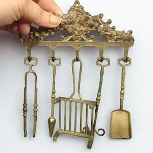 Op de afbeelding: Een antieke messing haardgereedschapsset met een bloemenopzet. De set bevat een tang, een pook, een kleine schop en een miniatuurrooster, allemaal hangend aan een decoratief rek. De gereedschappen zijn aangetast en tonen hun leeftijd.