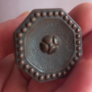 May include: An octagonal bronze-toned metal knob featuring a textured surface, a central four-petal floral motif, and a raised beaded border along the outer edge. The hardware has a matte finish and a geometric shape with eight distinct sides.