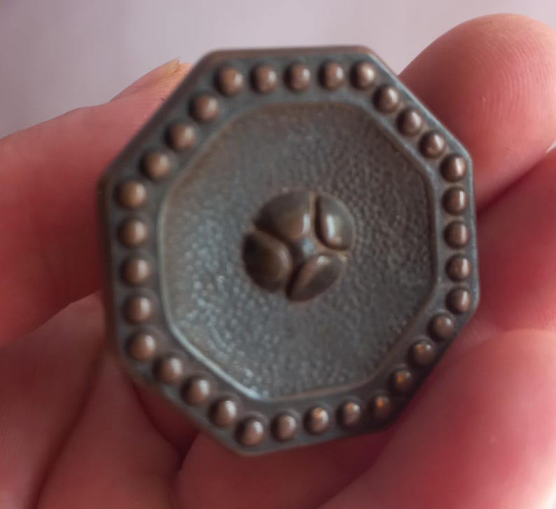 May include: An octagonal bronze-toned metal knob featuring a textured surface, a central four-petal floral motif, and a raised beaded border along the outer edge. The hardware has a matte finish and a geometric shape with eight distinct sides.