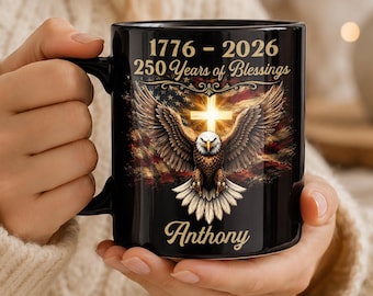 Op de afbeelding: Een zwarte keramische mok met een afbeelding van een Amerikaanse zeearend met gespreide vleugels, een stralend kruis en een vervaagde Amerikaanse vlag. De tekst luidt 1776 - 2026, 250 Years of Blessings en de naam Anthony in goud.