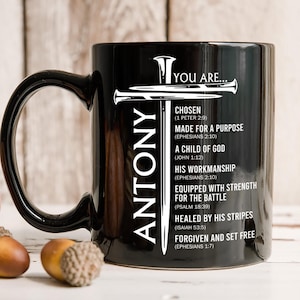 May include: Black ceramic mug with the name "ANTONY" in white vertical text. The mug features a white cross design and the words "YOU ARE..." and inspirational phrases like "CHOSEN" and "MADE FOR A PURPOSE".