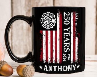 Op de afbeelding: Een zwarte keramische mok met een verweerd Amerikaans vlagontwerp in rood en wit. De afbeelding bevat een brandweerembleem, de tekst 250 YEARS, de jaartallen 1776-2026 en de naam ANTHONY gecentreerd onderaan tussen twee witte sterren.