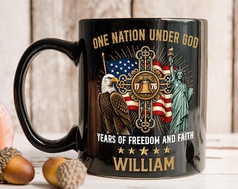 Op de afbeelding: Een zwarte keramische mok met een afbeelding met de tekst ONE NATION UNDER GOD en YEARS OF FREEDOM AND FAITH. Het ontwerp bevat een Amerikaanse zeearend, een Amerikaanse vlag, de Liberty Bell met 1776, het Vrijheidsbeeld en de naam WILLIAM.