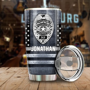 May include: A stainless steel tumbler with a black and white design featuring a correctional officer badge with the text "To Serve and Protect", "Corrections Officer", and "313". The tumbler also has a silver lid and the name "JONATHAN" printed on the side.