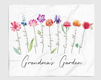 Op de afbeelding: Een wit deken met een bloemenontwerp en de tekst "Grandma's Garden". De bloemen zijn in verschillende kleuren en hebben namen eronder geschreven: Brady, Amabel, Emma, Anthony, Logan, Brady, Carol, Heather en Oriana.