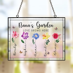 May include: A glass sign with a black frame and a white background. The sign says "Nana's Garden Love Grows Here" with five flowers and butterflies. The flowers are pink, purple, blue, yellow, and red. The names Brady, Sophia, Emma, David, and Olivia are written below the flowers.
