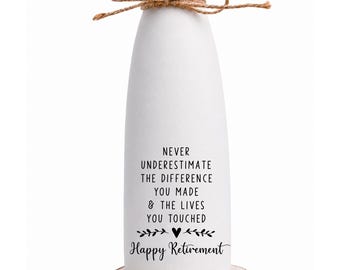 Op de afbeelding: Een witte keramische vaas met een uitlopende rand en een bruine touwstrik om de hals. De vaas is voorzien van zwarte tekst: NEVER UNDERESTIMATE THE DIFFERENCE YOU MADE & THE LIVES YOU TOUCHED Happy Retirement. Het staat op een houten schijf als voet.