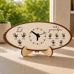 May include: Oval wooden clock with a bark-like edge. The clock face is light-colored wood, featuring a clock face, handprints, and the words "First dad" and "Now grandpa". Names are printed below the handprints. The clock is supported by a wooden stand.