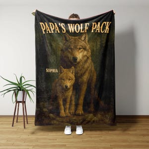 May include: A black blanket featuring a detailed illustration of two wolves. The larger wolf stands behind a smaller one. The text "PAPA'S WOLF PACK" is at the top, with "SOPHIA" below. The blanket has a dark green and brown background.