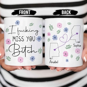 May include: White ceramic mug with the text "I fucking MISS YOU Bitch" in black script, decorated with pink and blue floral accents. The back of the mug features state outlines, names, and a connecting heart design.