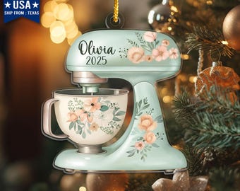 May include: A light blue stand mixer ornament with floral accents and the name "Olivia" and the year "2025". The ornament is decorated with peach and green floral designs. The text "MADE IN USA" and "SHIP FROM: TEXAS" is visible.