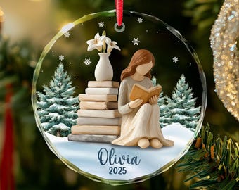 May include: A clear, round Christmas ornament with a red ribbon. Inside, a figure reads a book near a stack of books and a vase of flowers. The ornament has a winter scene with snowflakes, pine trees, and the name "Olivia" and the year "2025".