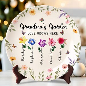 May include: A decorative ceramic plate with the words "Grandma's Garden" and "Love Grows Here." The plate features colorful flowers, butterflies, and names. The plate is displayed on a brown stand.