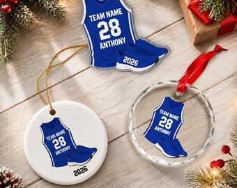 Op de afbeelding: Drie kerstornamenten met een blauw basketbalshirt met de tekst TEAM NAME, 28 en ANTHONY, gecombineerd met blauwe sportschoenen. Eén ornament heeft de vorm van een shirt, één is een witte cirkel met 2026 en één is een doorzichtige glazen cirkel.