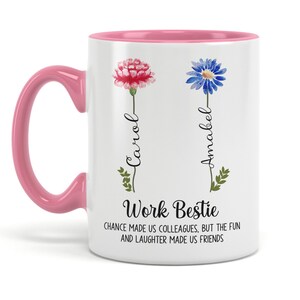 May include: White ceramic mug with a pink rim and handle. The mug features a floral design with two flowers, a pink carnation and a blue cornflower. The text "Carol" and "Amabel" are written vertically next to the flowers. The text "Work Bestie" is written in black at the bottom of the mug, along with the text "CHANCE MADE US COLLEAGUES, BUT THE FUN AND LAUGHTER MADE US FRIENDS."