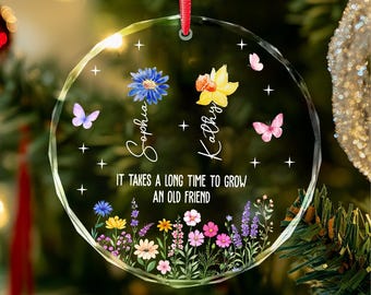 Op de afbeelding: Een rond, helder, gefacetteerd glazen ornament dat aan een rood lint hangt. Het toont een blauwe bloem, een gele bloem, vlinders en een veld met wilde bloemen. De tekst luidt: Sophia, Kathy, IT TAKES A LONG TIME TO GROW AN OLD FRIEND.