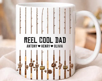 Op de afbeelding: Een witte keramische mok met een herhalend patroon van bruine hengels en molens. De tekst REEL COOL DAD is in het zwart gedrukt, gevolgd door de namen ANTONY, HENRY en OLIVIA gescheiden door zwarte hartjes in het midden van de mok.