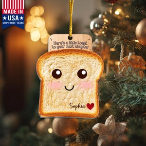 May include: A novelty Christmas tree decoration in the shape of a slice of toast with a smiling face, pink cheeks, and the name "Sophia" with a heart. The ornament reads "Here's a little toast to your next chapter."