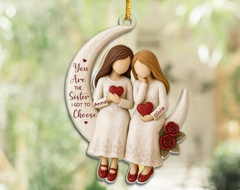 Op de afbeelding: Een hangend ornament met twee figuren met bruin en blond haar die op een halve maan zitten. De figuren dragen lange crèmekleurige jurken en houden rode harten vast. Tekst: You Are the Sister I Got to Choose, met de namen Anna en Olivia.