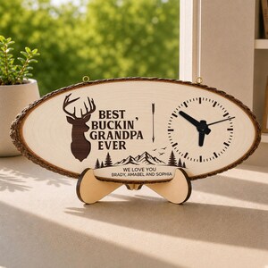 May include: Oval wooden clock with a natural bark edge. The clock face features a deer silhouette, the text "BEST BUCKIN' GRANDPA EVER", an arrow, and a clock face. The base reads "WE LOVE YOU, Brady, Amabel and Sophia."