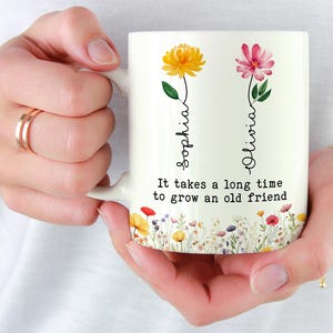 May include: White ceramic mug featuring floral designs and the names "Sophia" and "Olivia." The mug has a floral border and the text "It takes a long time to grow an old friend." The mug is held by a person.