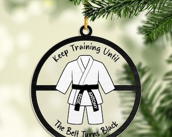 May include: A round, black and white ornament with a karate uniform illustration. The text reads "Keep Training Until The Belt Turns Black" and includes the name "Antony" and the year "2025". The ornament hangs from a gold string.