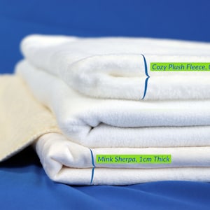May include: A stack of white fabric blankets. The top blanket is labeled "Cozy Plush Fleece, 0.6cm Thick". The bottom blanket is labeled "Mink Sherpa, 1cm Thick".
