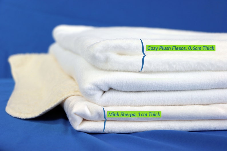 May include: A stack of white fabric blankets. The top blanket is labeled "Cozy Plush Fleece, 0.6cm Thick". The bottom blanket is labeled "Mink Sherpa, 1cm Thick".