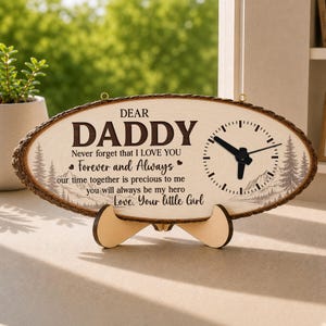 May include: Oval wooden clock with a bark-like edge, featuring the text "DEAR DADDY" and a heartfelt message. The clock face includes a clock and a mountain and tree design. It stands on a wooden stand.