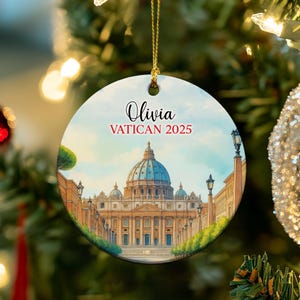 May include: Circular Christmas tree decoration with a watercolour depiction of St Peter's Basilica in Vatican City. The ornament is inscribed with the name "Olivia" and the text "VATICAN 2025". It is suspended by a gold-coloured string.