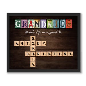 May include: A framed print with a brown wood background featuring the word "GRANDKIDS" spelled out in colorful wooden blocks. Below the word is the phrase "make life more grand" in white script. The names "ANTONY" and "CHRISTINA" are spelled out in a crossword puzzle format using wooden letter tiles.