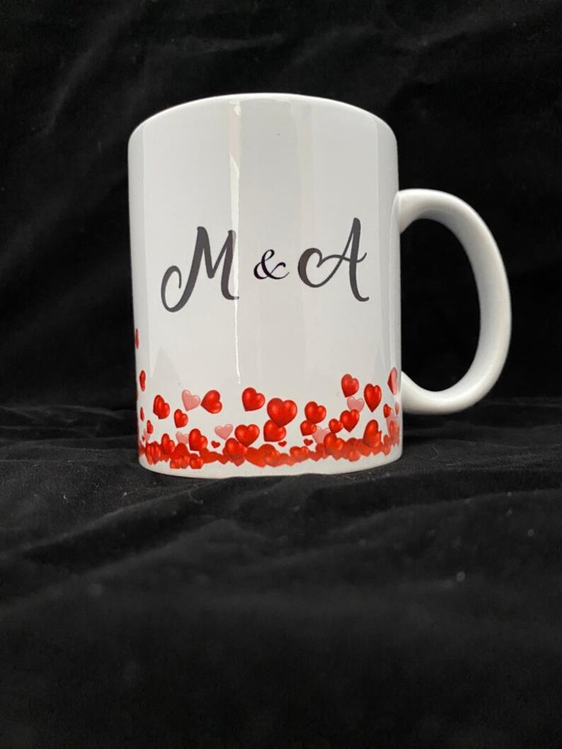May include: White ceramic mug with a black inscription "M & A" and a border of red hearts.