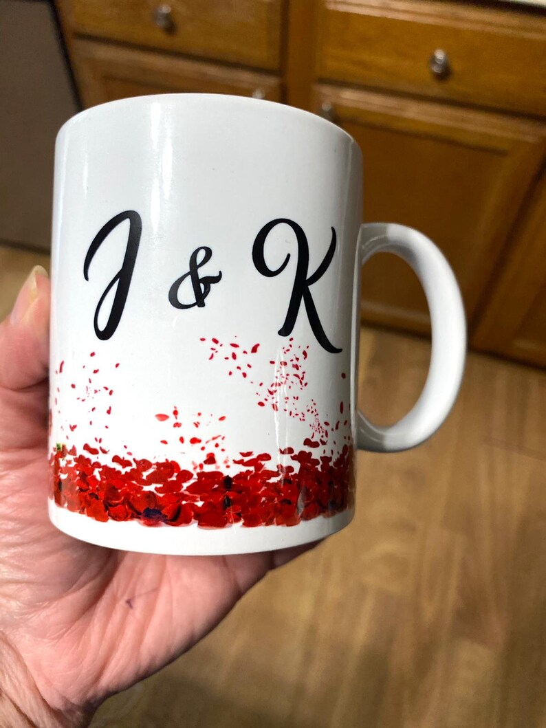 May include: White ceramic mug with a black design of "J & K" and a red and white confetti pattern around the bottom.