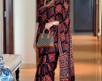 Op de afbeelding: Een vrouw draagt een sari met patronen in rood, zwart en blauw, met een geometrisch ontwerp. Ze draagt een bruine handtas met een geometrisch patroon en gouden sieraden. De sari is een traditioneel kledingstuk.