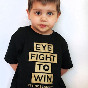 May include: A black short-sleeved t-shirt featuring gold block lettering that reads EYE FIGHT TO WIN, with the text RETINOBLASTOMA WARRIOR printed in smaller gold letters underneath the main graphic on the front of the garment.